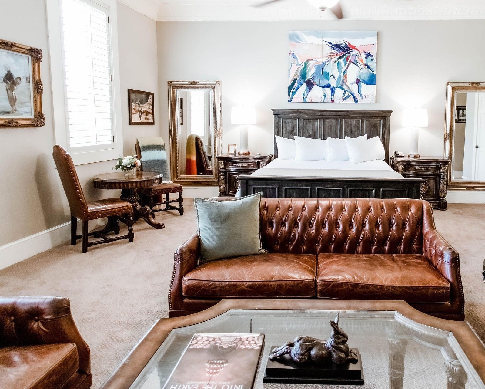 Front view of a well decorated bedroom with a leather sofa at its feet accompanied by  a glass table,a wooden table with a  chair, two mirrors on either corner of the room and drawers at either side of the bed. and art works on the wall - The Grand Hotel &