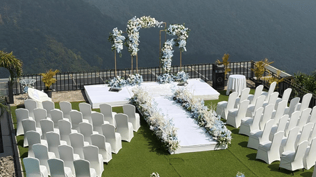 Outdoor wedding ceremony setup at Polo Resort Cherrapunji with white chairs, floral arch and aisle on grass overlooking misty green mountains.