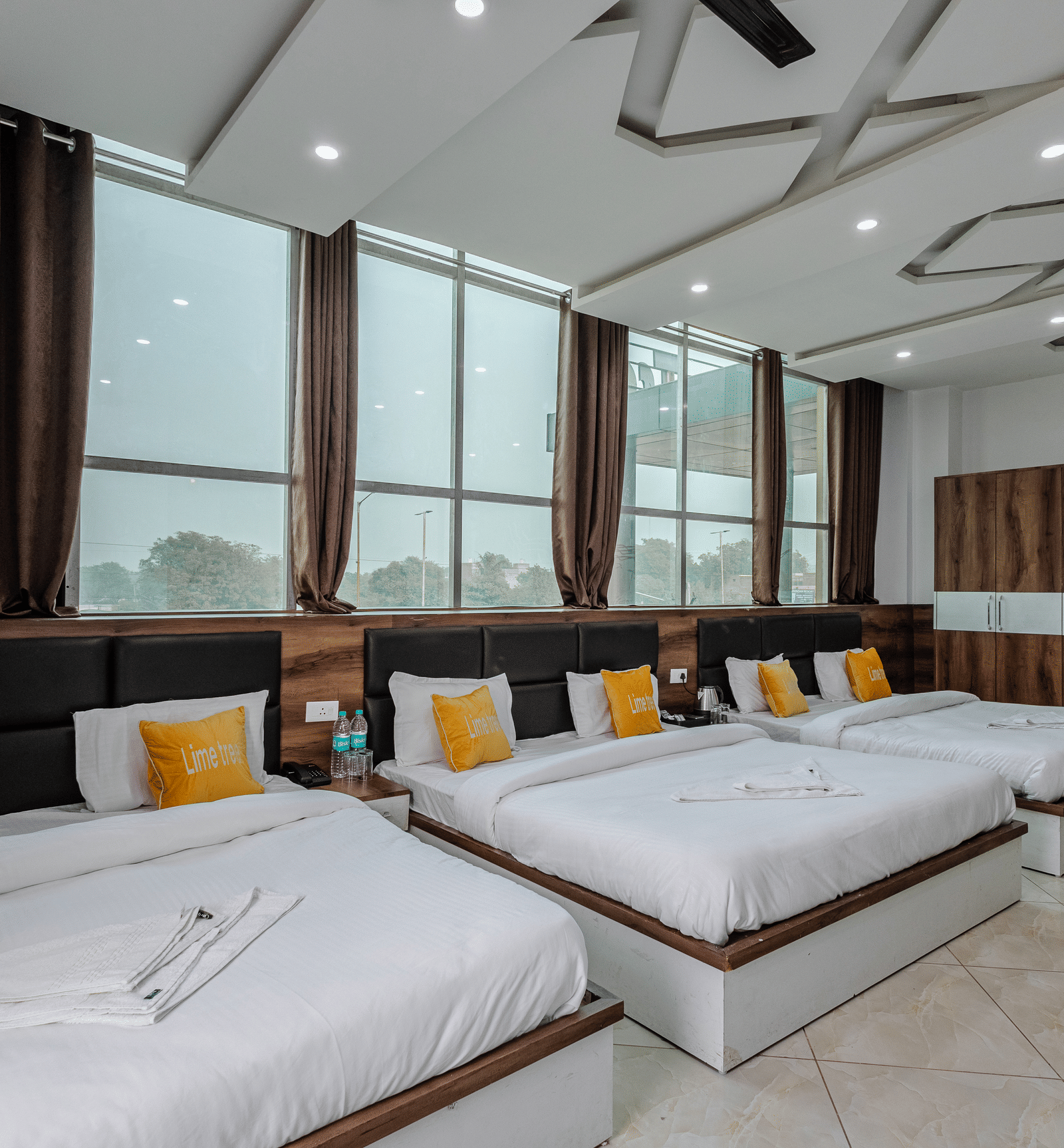 A room with three double beds lined up in a room with huge windows behind it at Lime Tree Hotels and Banquet, Vrindavan.