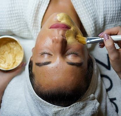 A close-up of a woman receiving a facial treatment, with a paste being applied to her face by a brush.