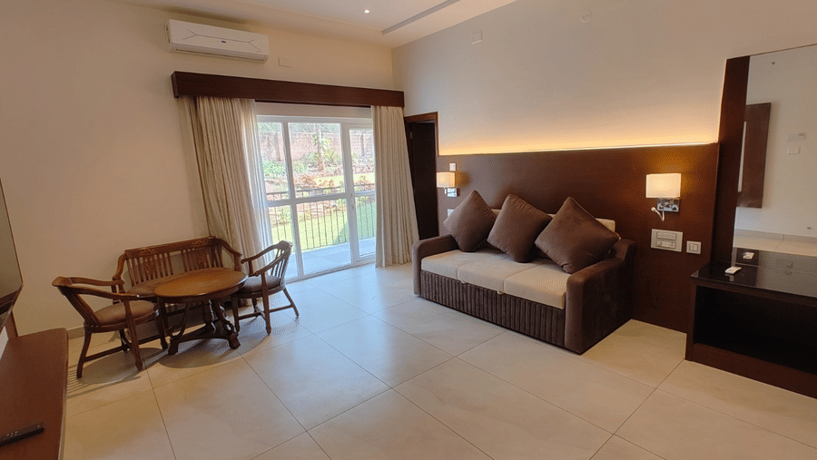 A spacious hotel room featuring a double bed, a brown sofa, a small dining table with chairs, and a large window with curtains | Lords Sahyadri Resort, Shivamogga