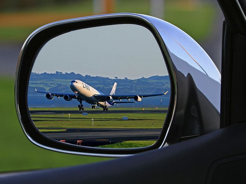 An aeroplane ascends from a runway, its image reflected in a car's side mirror, showing the aircraft against a landscape.
