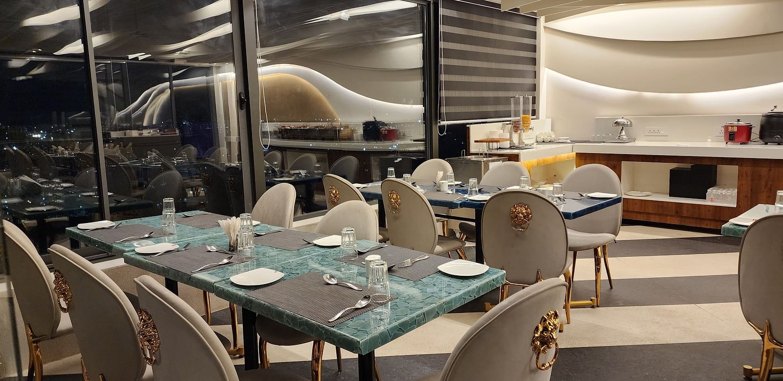 An elegant dining area with white and gold chairs and set tables, located beside large windows offering a view outside.