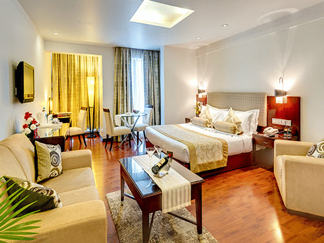 Wide view of a Premium Room at Hotel Hindusthan International Select Bengaluru with a king bed, cream sofas, wooden flooring, coffee table, and skylight.
