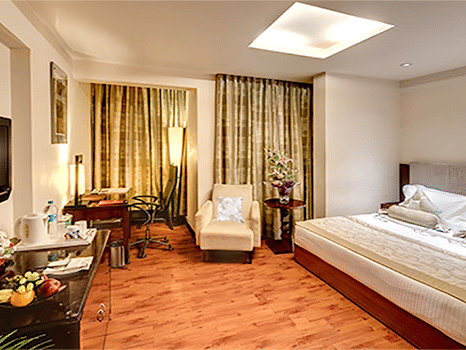View of a Premium Room at Hotel Hindusthan International Select Bengaluru with a king bed, wall-mounted TV, armchair, skylight, and warm wooden flooring.