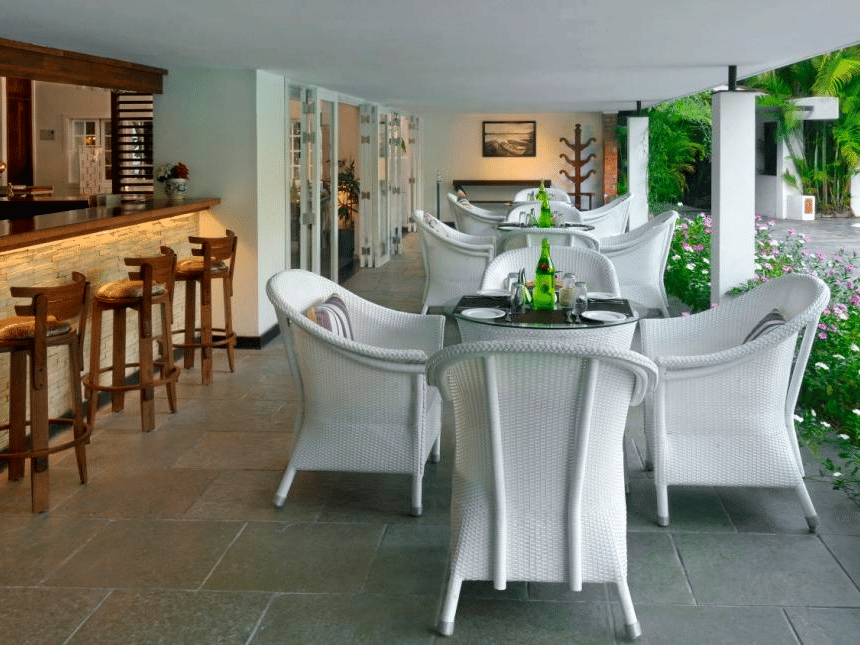 Modern covered patio with bar and white tables and chairs at Amanvana Spa Resort Coorg overlooking lush green surroundings.