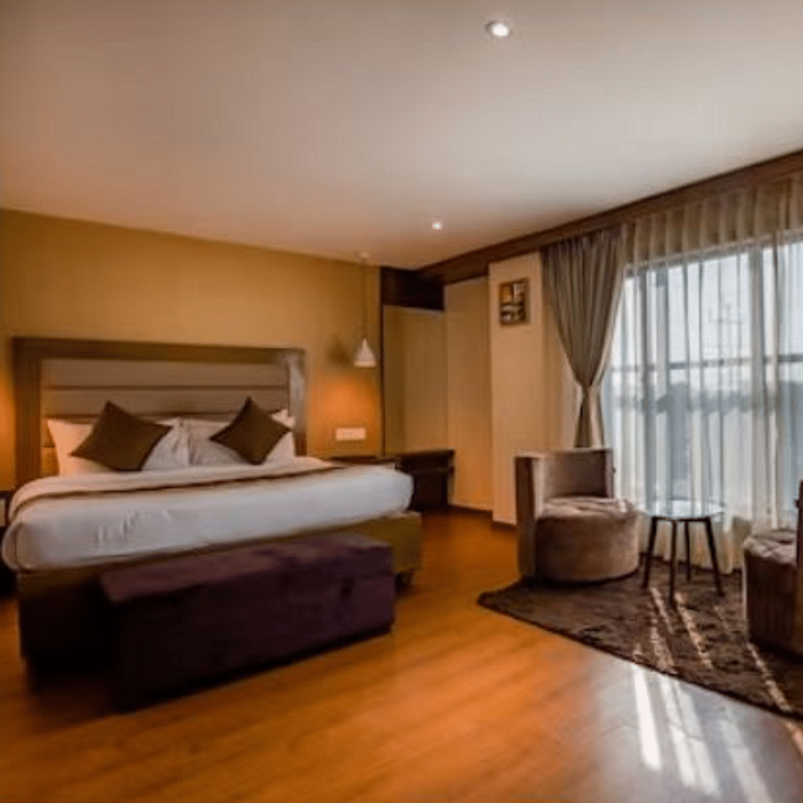 A premium hotel room with a large bed and windows that extend from the ceiling to the floor, with curtains pulled to the side, highlighting the two chairs and table at Yashshree The Imperial Inn, Siliguri.