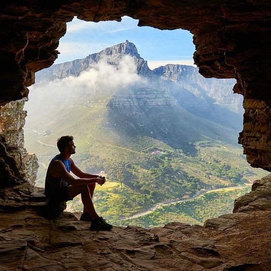 Photo of a man sitting inside a cave with hills in the background. 