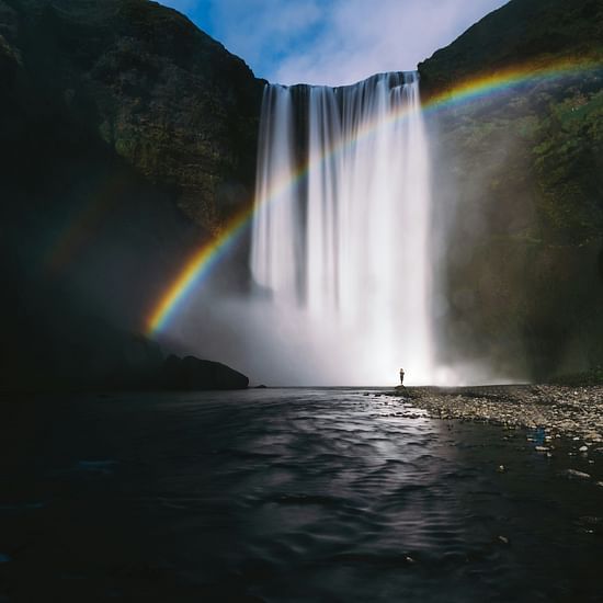 A person standing at the edge looking at the waterfalls with a rainbow in front of the falls