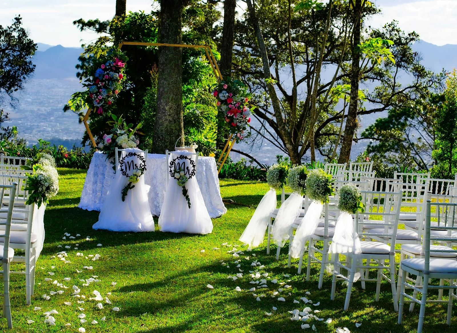 A wedding setup on a green lawn with chairs facing a decorated archway under trees, suggesting an outdoor ceremony.