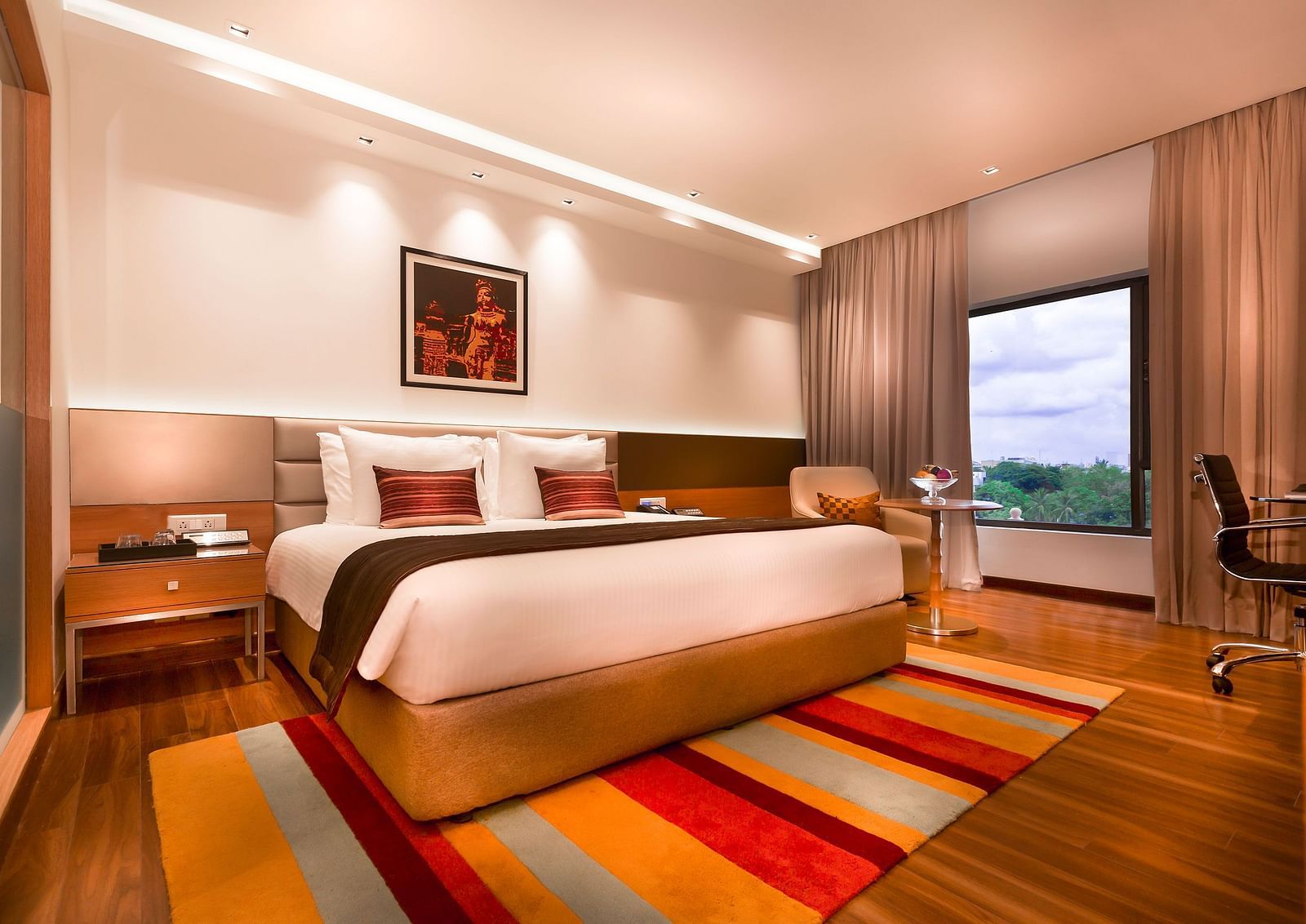 A modern hotel room featuring a king size bed, warm lighting, wooden flooring, a striped rug, and a large window with a view - Grand Chennai by GRT Hotels