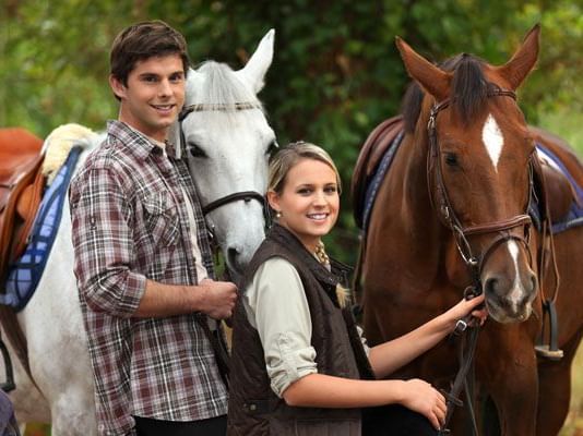 A man and a woman posing with a white and brown horse in an outdoor setting.