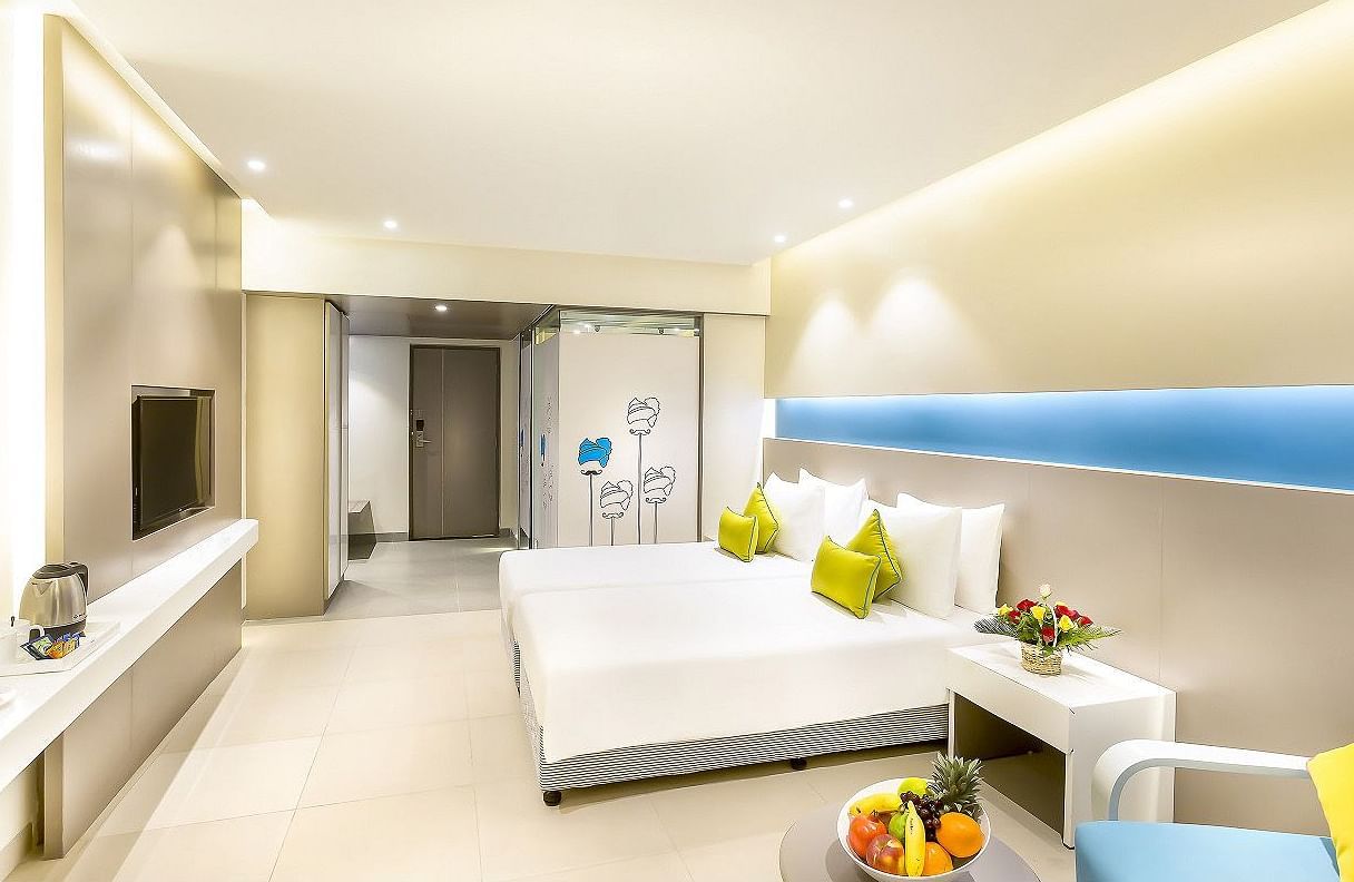 A spacious room features a large bed with a headboard stretching across the wall. Cushion chairs sit beside a wooden bedside table, and a wall-mounted television is positioned opposite the bed