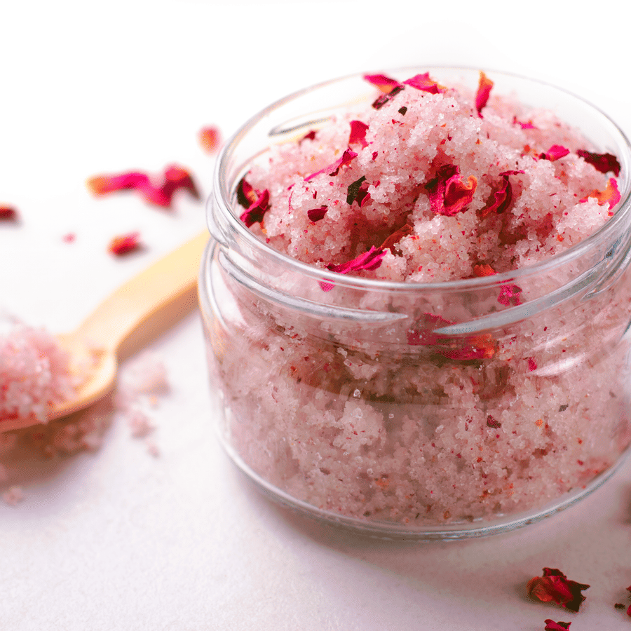 Jar of pink body scrub with petals and a wooden spoon on the side.