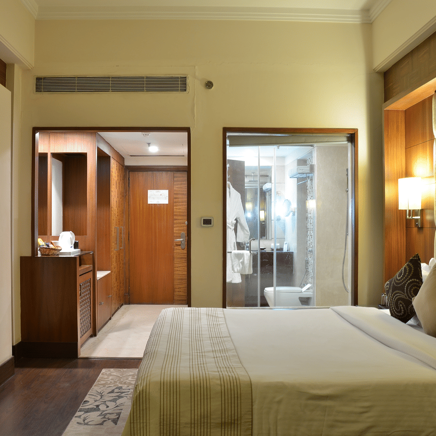 Premium Room at The Bristol Hotel, Gurgaon with a king-size bed, wall-mounted TV, a wooden wardrobe area, and a glass-enclosed bathroom with a walk-in shower.