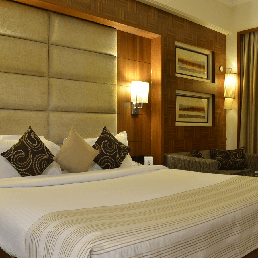 Premium Room at The Bristol Hotel, Gurgaon with a king-size bed, tall padded headboard, brown patterned cushions, framed wall art, wall sconces, and a sofa.