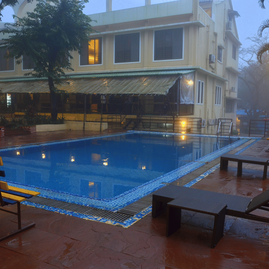 A swimming pool reflects the soft glow of nearby lights, with lounge chairs placed along the wet deck and a building partially veiled in mist in the background | Zara's Resort, Khandala