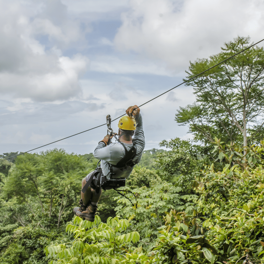 A close-up of a man ziplining through a lush green forest, wearing a helmet and harness, with an excited expression as he glides above the treetops.