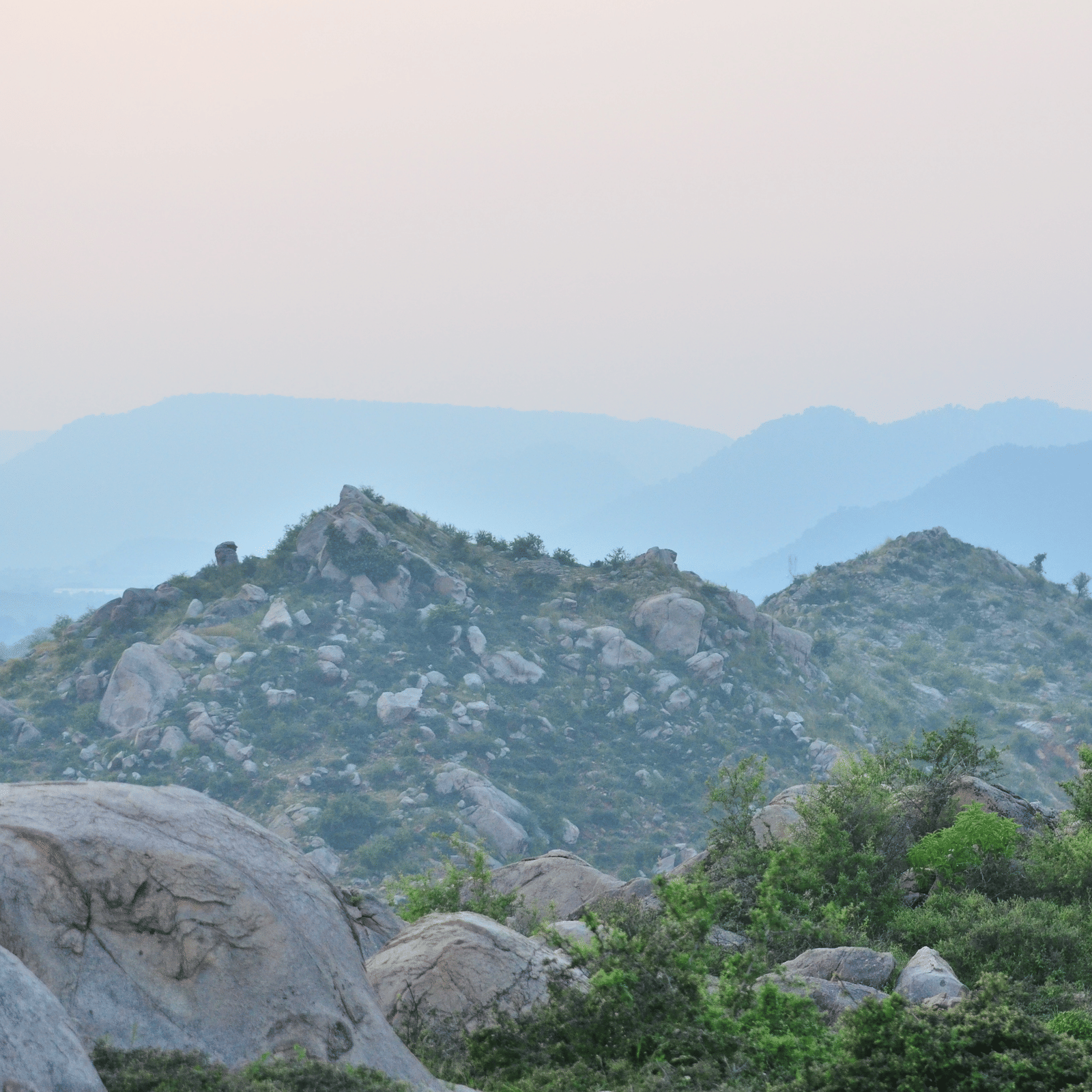 Hazy view of rocky hilltops covered in scattered shrubs and boulders, with layered mountain ridges in the background during dawn or dusk.