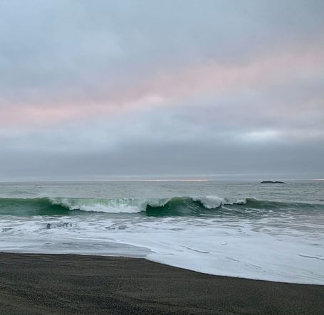 A wave curls and breaks onto a dark sandy beach under a cloudy sky with a faint hint of light.