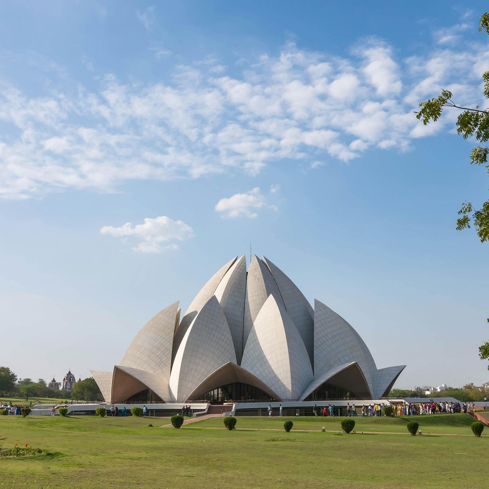The Lotus Temple, a modern architectural marvel shaped like a lotus flower with white petal-like structures, stands in a green landscape under a blue sky with scattered clouds.