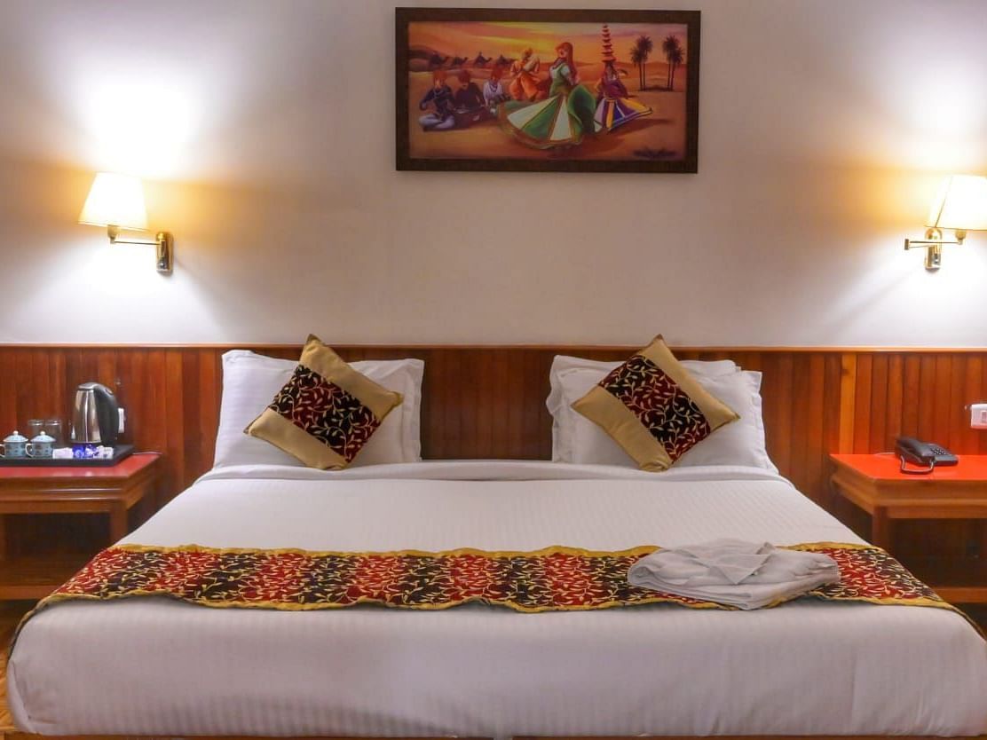 Warmly lit hotel room featuring a double bed with patterned cushions and runner, bedside tables with lamps, a tea kettle setup, and a traditional painting above the headboard - Sumitel Chiminda, Pelling.