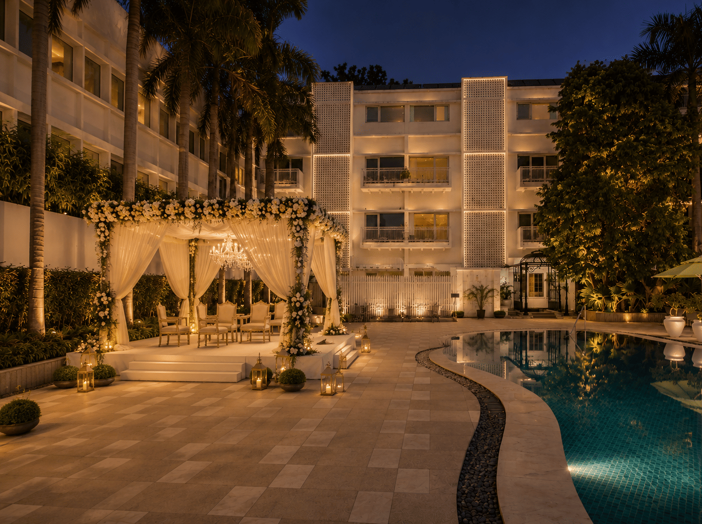 Night view of an illuminated outdoor cabana with white drapes set beside a swimming pool at a hotel courtyard - The Claridges New Delhi
