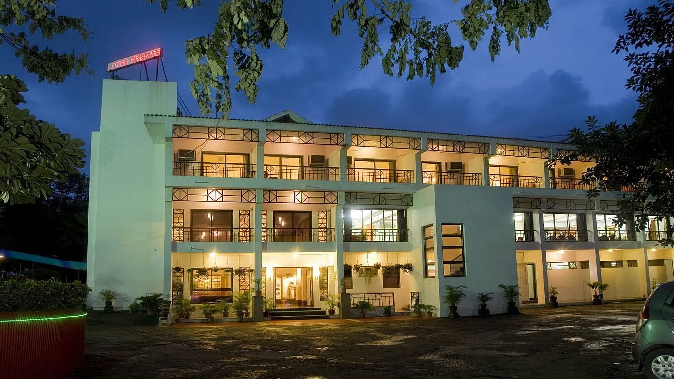 Exterior view of a well-lit two-storey building at dusk, with large windows and a visible red sign on the rooftop, surrounded by trees and parked vehicles.