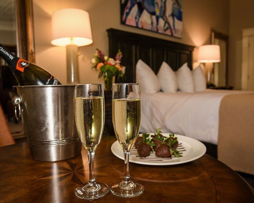 Front view of two champagne glasses filled with champagne , a steel ice bucket and a plate of food on a table in dimly lit room with a neatly made bed and lamps on either side of the bed