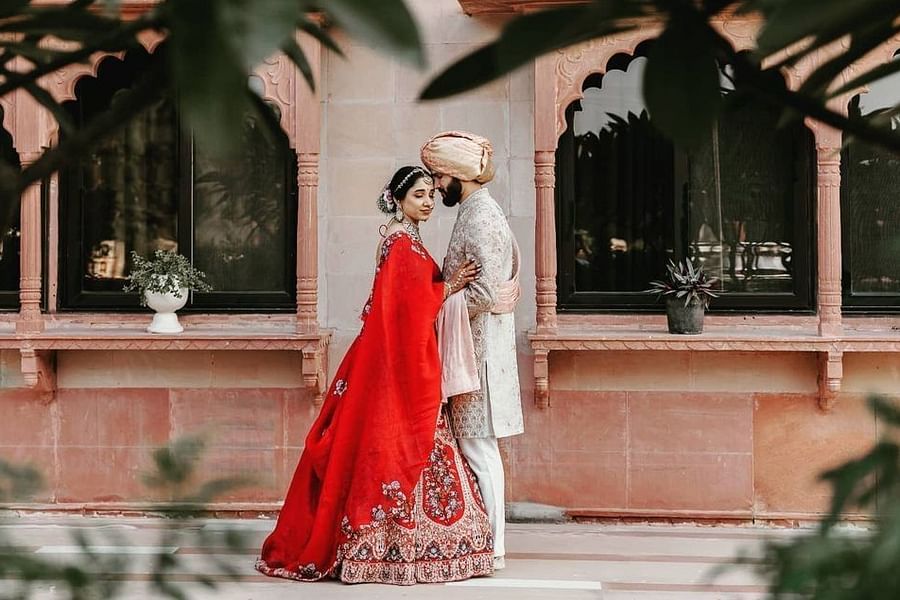 A bride and groom embrace outdoors, framed by foliage with traditional architecture behind them - Umaid Palace- Best Luxury Heritage Resort Near Jaipur.  
