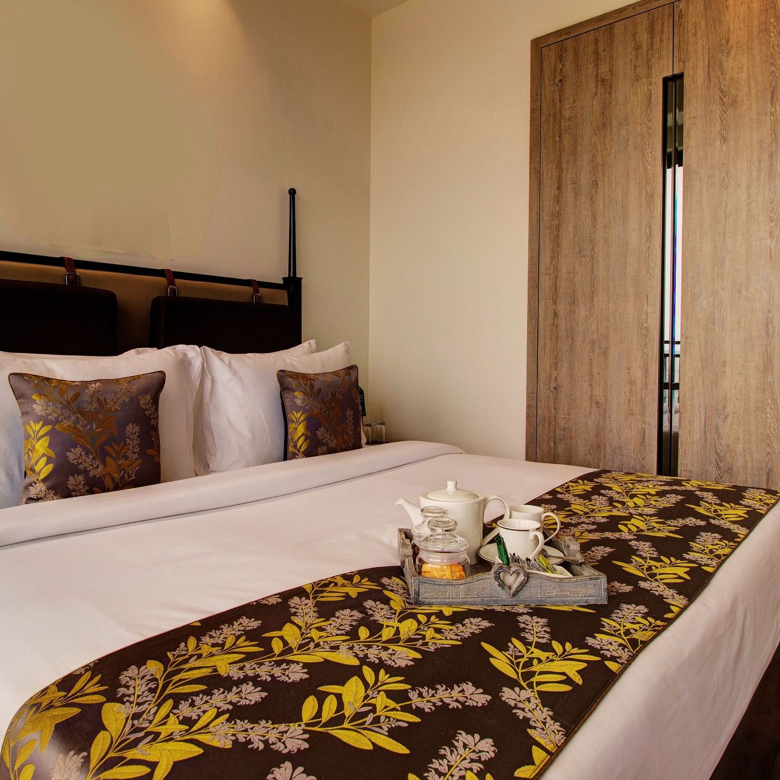 A plush bed on with pillows and cushions and a tray containing cutlery for hot beverages sitting on the bed runner with a large closet in the background