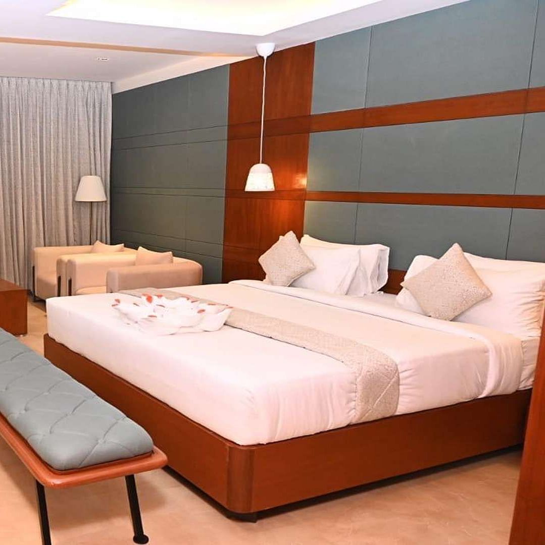 A Executive Suite at Dhanalakshmi Srinivasan Hotels, Perambalur featuring a neatly made bed with white linens and pillows, a wooden headboard, and a desk with a chair.
