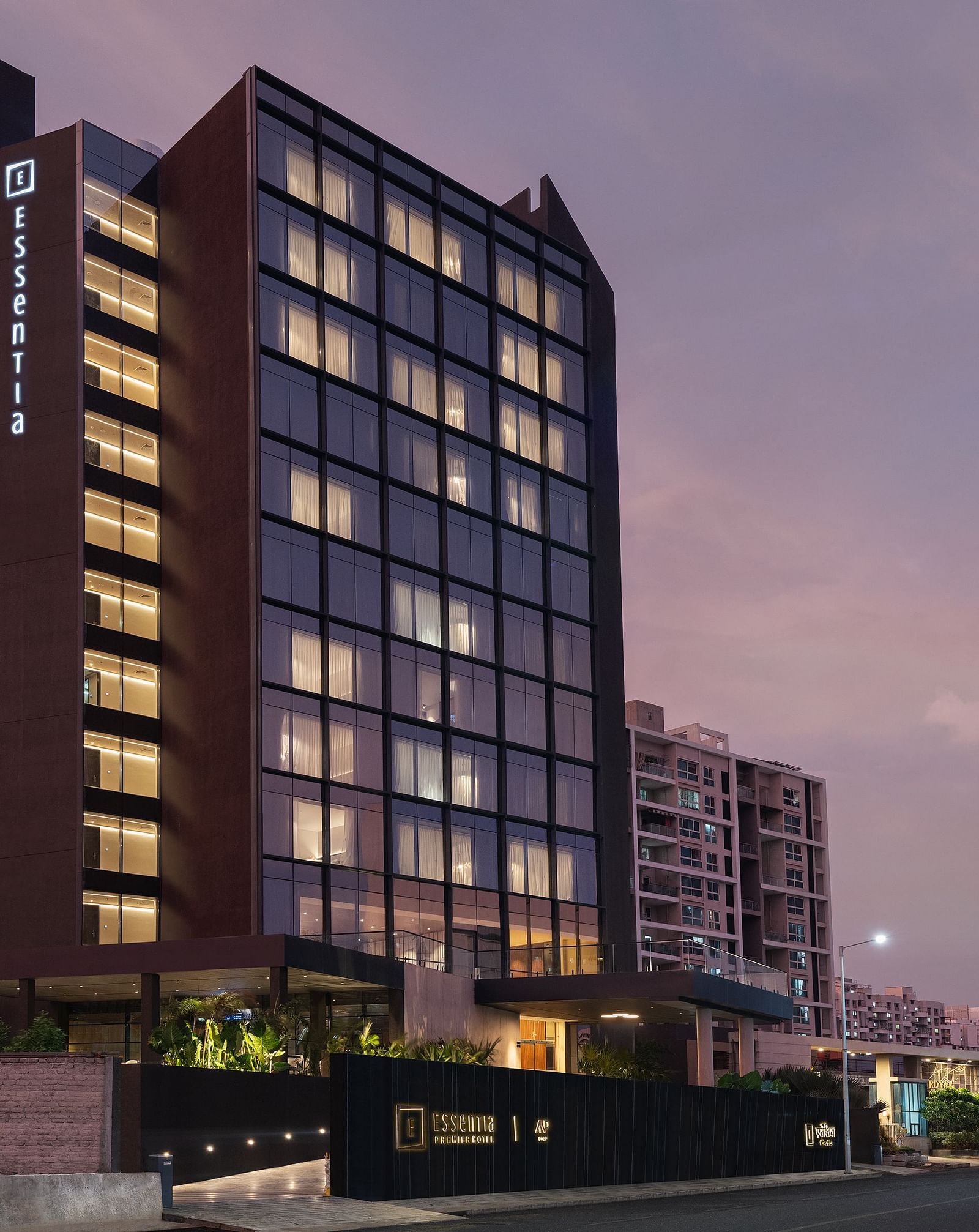 Facade of Essentia Premiere, Pune with lights on during twilight.