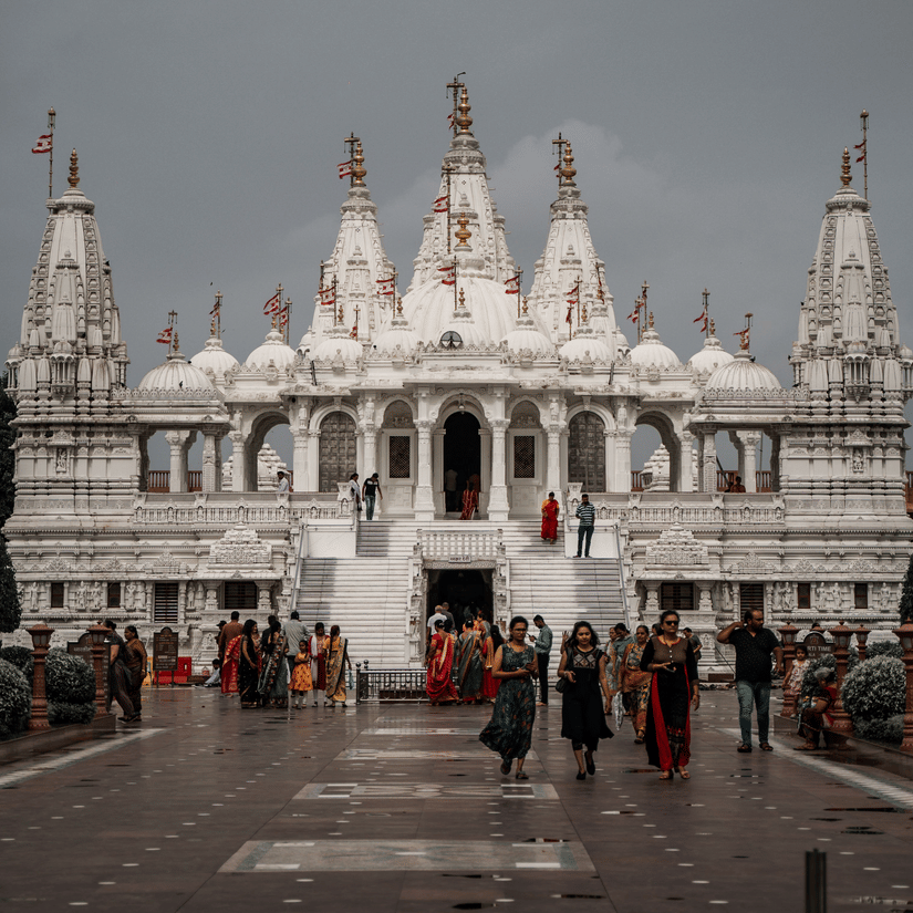 The BAPS Shree Swaminarayan Temple in Jamnagar with people walking in the path in front of it under a cloudy sky.