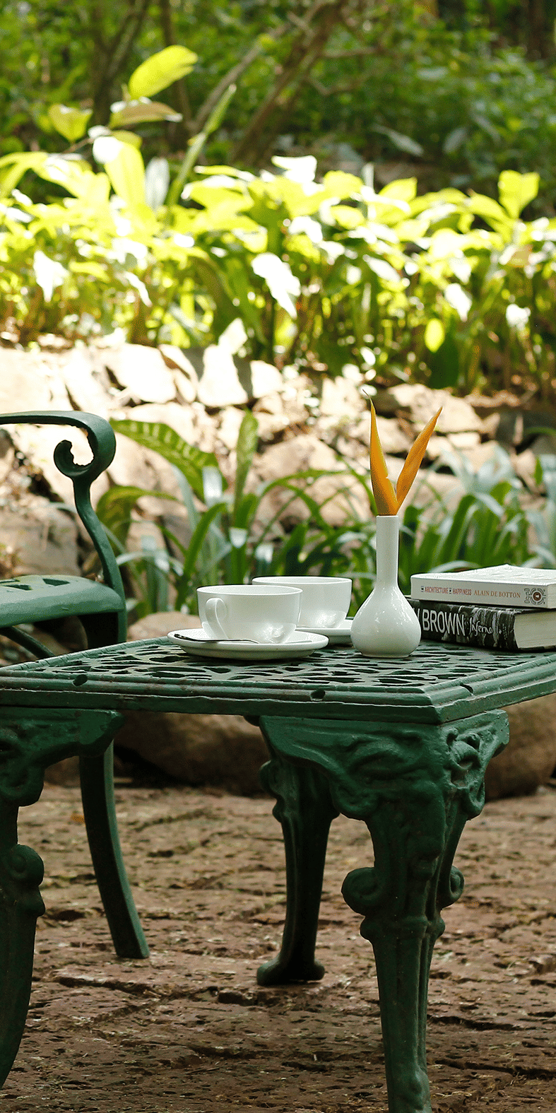 A green metal garden chair and table with a book, coffee mugs, and a small vase in a peaceful garden at Amanvana Spa Resort, Coorg – Luxury Resort.