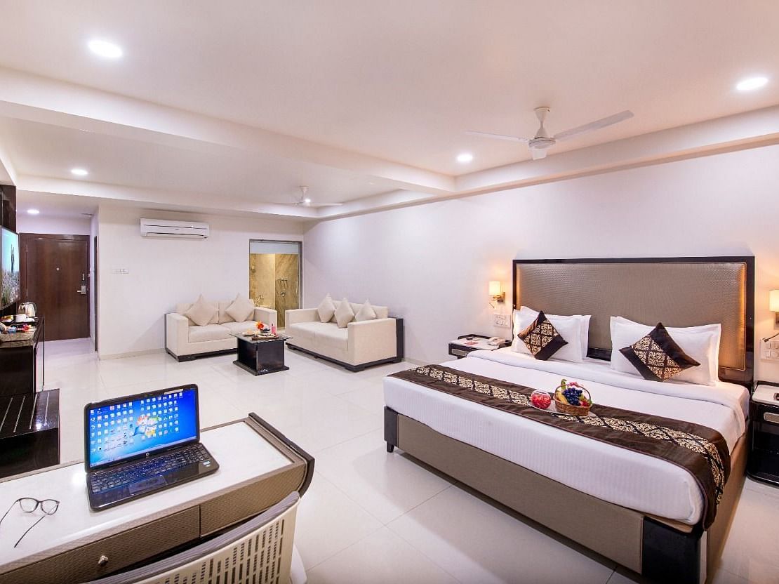 A comfortable hotel suite offers a king size bed, a separate seating area, a functional desk with a laptop, and ample natural light.