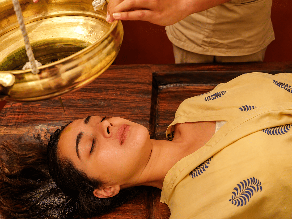 A woman experiencing a relaxing, traditional Ayurvedic wellness treatment at Neemrana’s Glasshouse on The Ganges, Rishikesh, with oil poured on her head from a brass vessel.