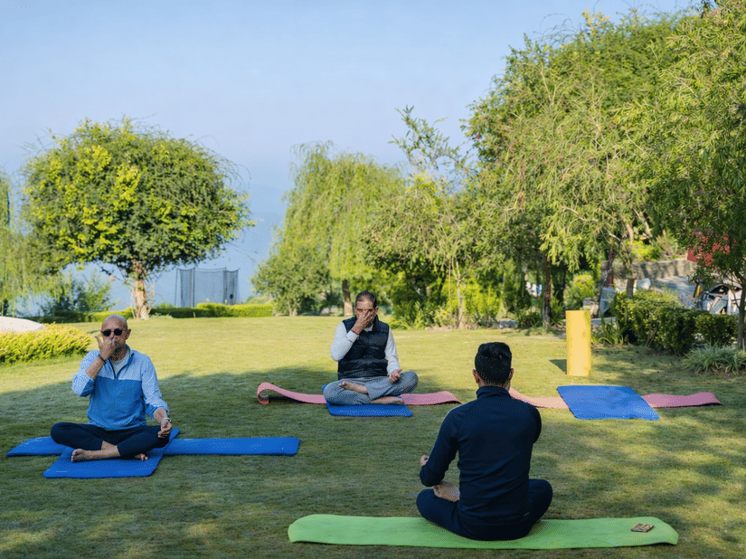 A group of people sitting on yoga mats outdoors in a park or garden, likely meditating or doing yoga  - Manor Sports & Wellness Hotel