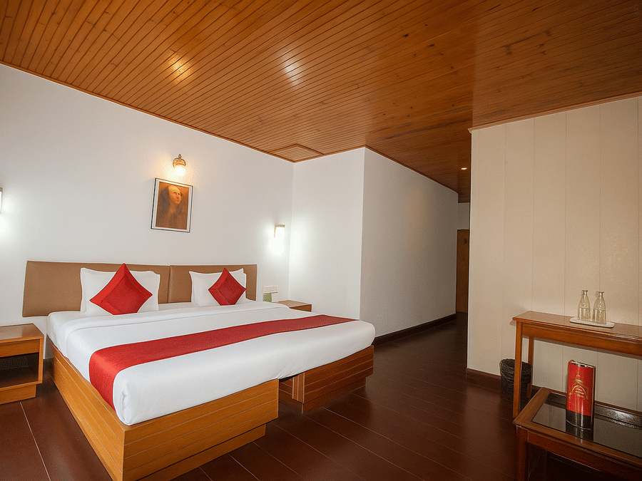 A room at Central Hotels featuring a king bed with red and white bedding wooden ceiling a work desk with chair wall lamps and dark hardwood flooring in a bright setting.