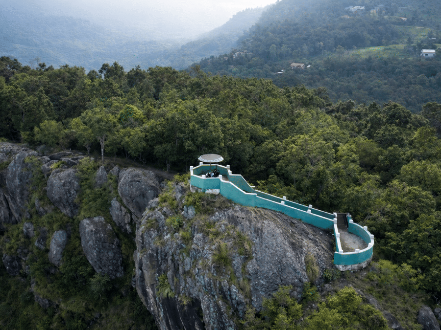 Aerial view of Lambs Rock in Coonoor overlooking lush green valleys and misty hills, with a viewing platform perched dramatically along the rocky cliff edge.