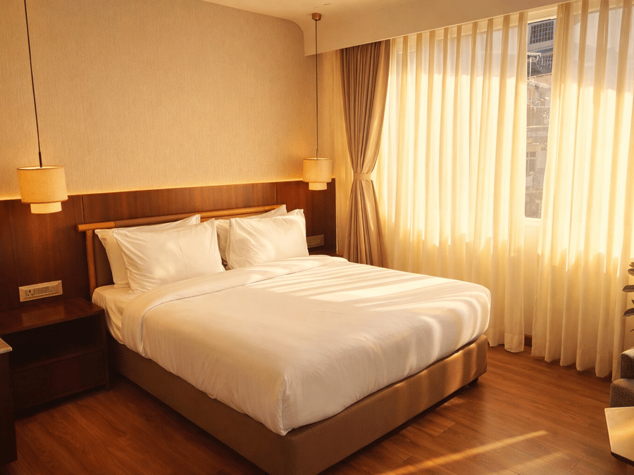 Bedroom of Pride Premier Darjeeling featuring a double size bed, bedside table with lamps on either side, and a large window with curtains drawn.