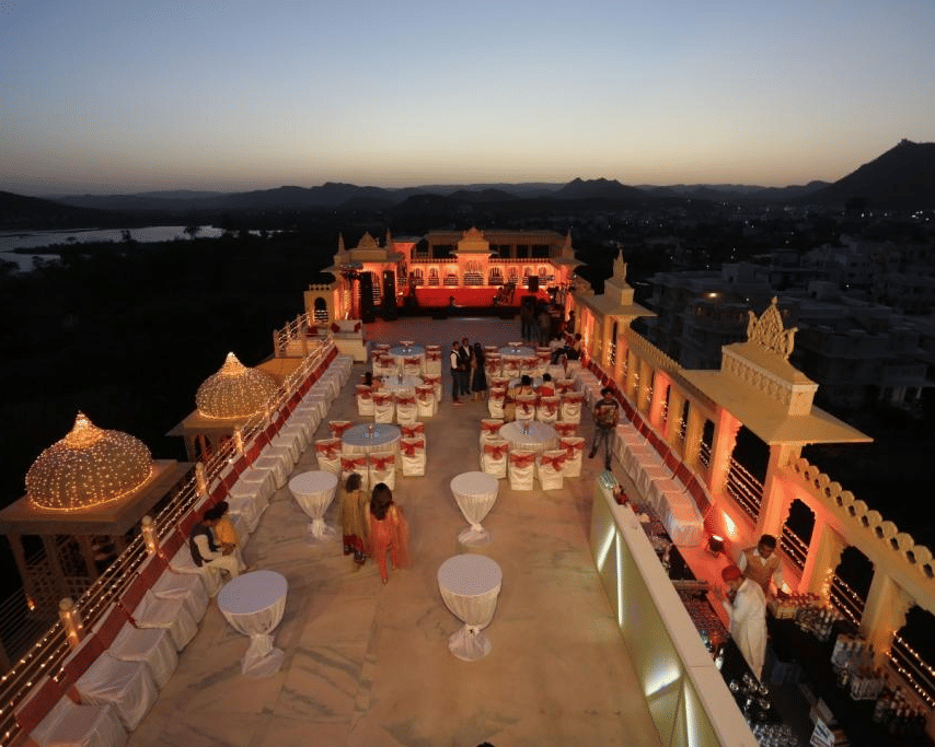 Aerial view of the top deck venue at Chunda Palace, Udaipur, during twilight, featuring round banquet tables, a bar counter setup, and small illuminated domes bordering the terrace.