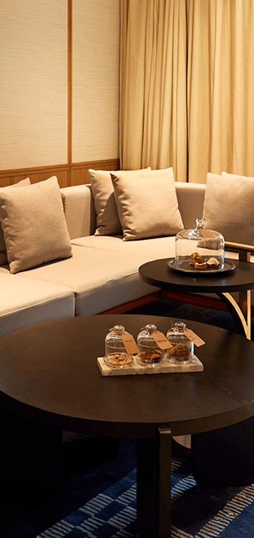 Modern hotel suite living area with a large sectional sofa, coffee tables, blue patterned rug, and a peek into the separate bedroom.