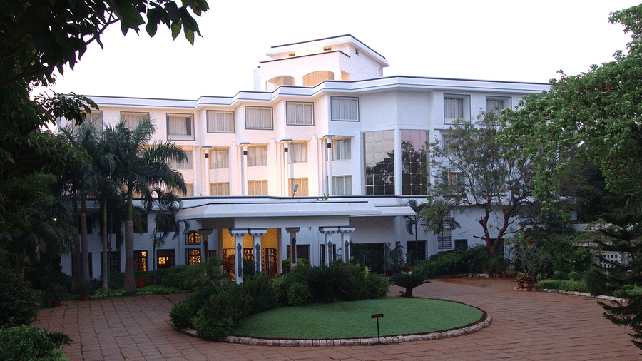 Facade view of Sangam Hotel, Thanjavur, with a pathway, a manicured garden and trees in view - Thanjavur Hotel Contact