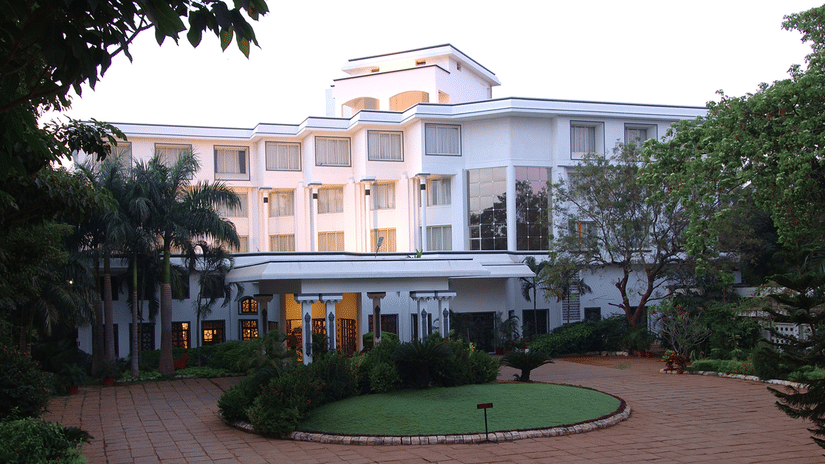 Facade view of Sangam Hotel, Thanjavur, with a pathway, a manicured garden and trees in view - Thanjavur Hotel Contact