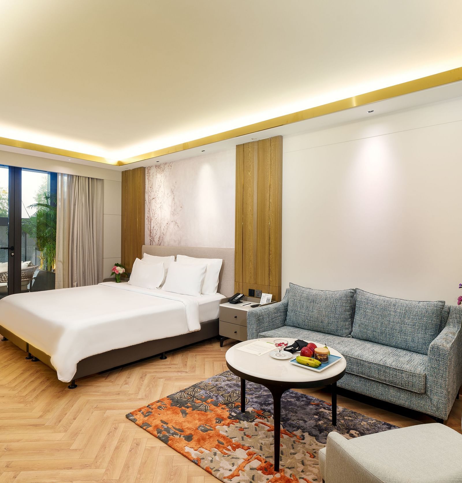 A bright and airy hotel room at Nirvana Luxury Hotel, Ludhiana, with a bed, a comfy blue sofa, a round table with fruit, and a colourful rug on the wooden floor.