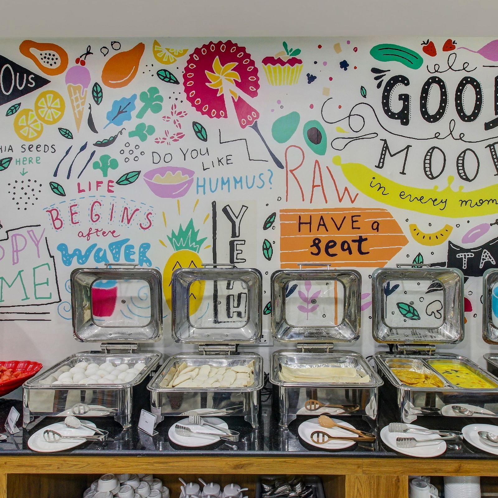 A hotel buffet corner with a wall mural saying Good Mood Bon Appétit and various other cheerful phrases alongside a spread of breakfast items ready for guests.