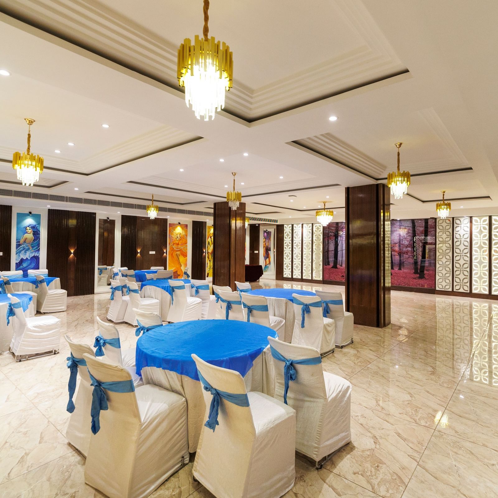 Banquet hall at Inde Reva Resorts Rishhikesh set up with round tables, white chairs, blue covers, and ceiling chandeliers.