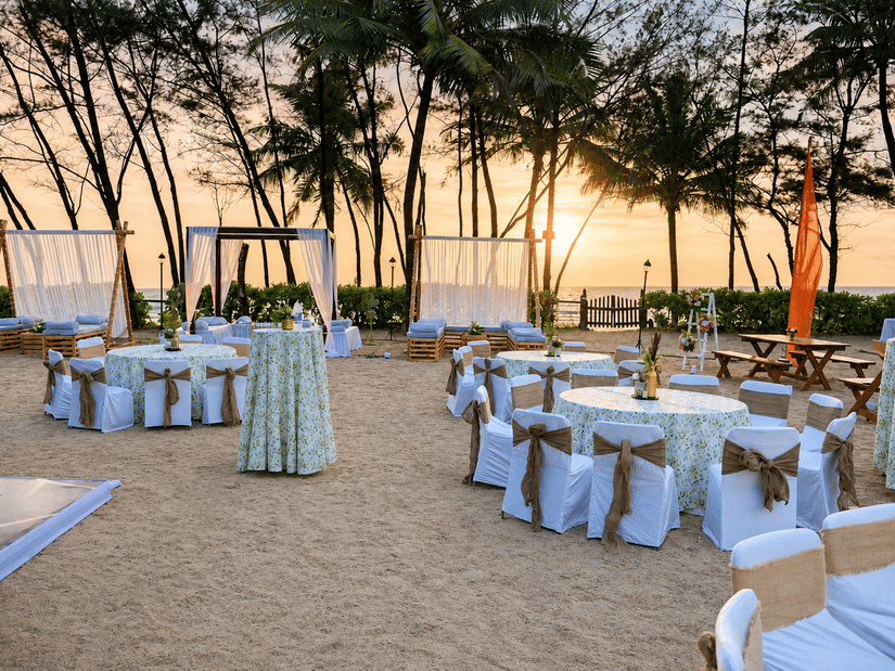 Outdoor beachfront event setup with decorated dining tables under palm trees at sunset at Kenilworth Resort & Spa, Goa.