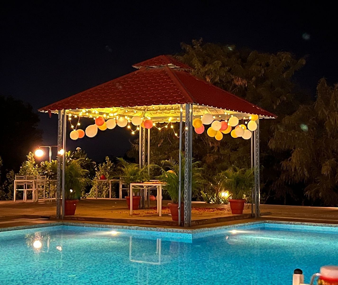 A nighttime shot of a gazebo with a red roof, decorated with lights and balloons, next to a brightly lit swimming pool - Bamboo Saa Mulberry Resort, Pushkar